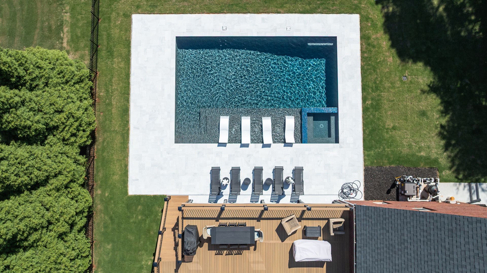 Overhead view of a backyard pool with white lounge chairs and a wooden deck beside a house.