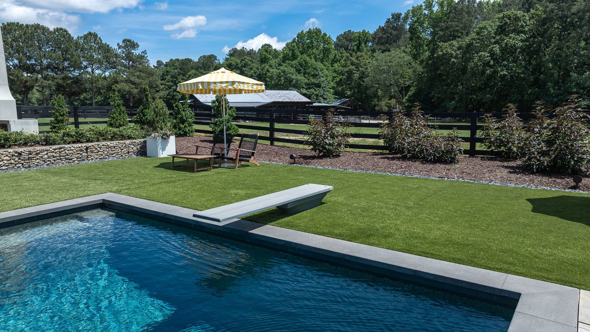 Backyard pool with manicured lawn, patio umbrella, lounge chairs, and stone fence under a sunny sky