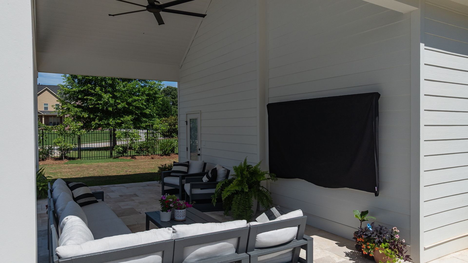Covered patio with gray outdoor sofa, black wall-mounted TV, and potted plants beside a white house wall