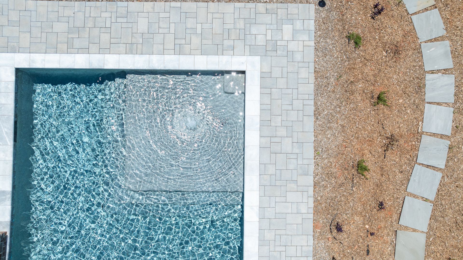 Aerial view of a pool with light stone deck beside a curved gravel path and sparse landscaping.