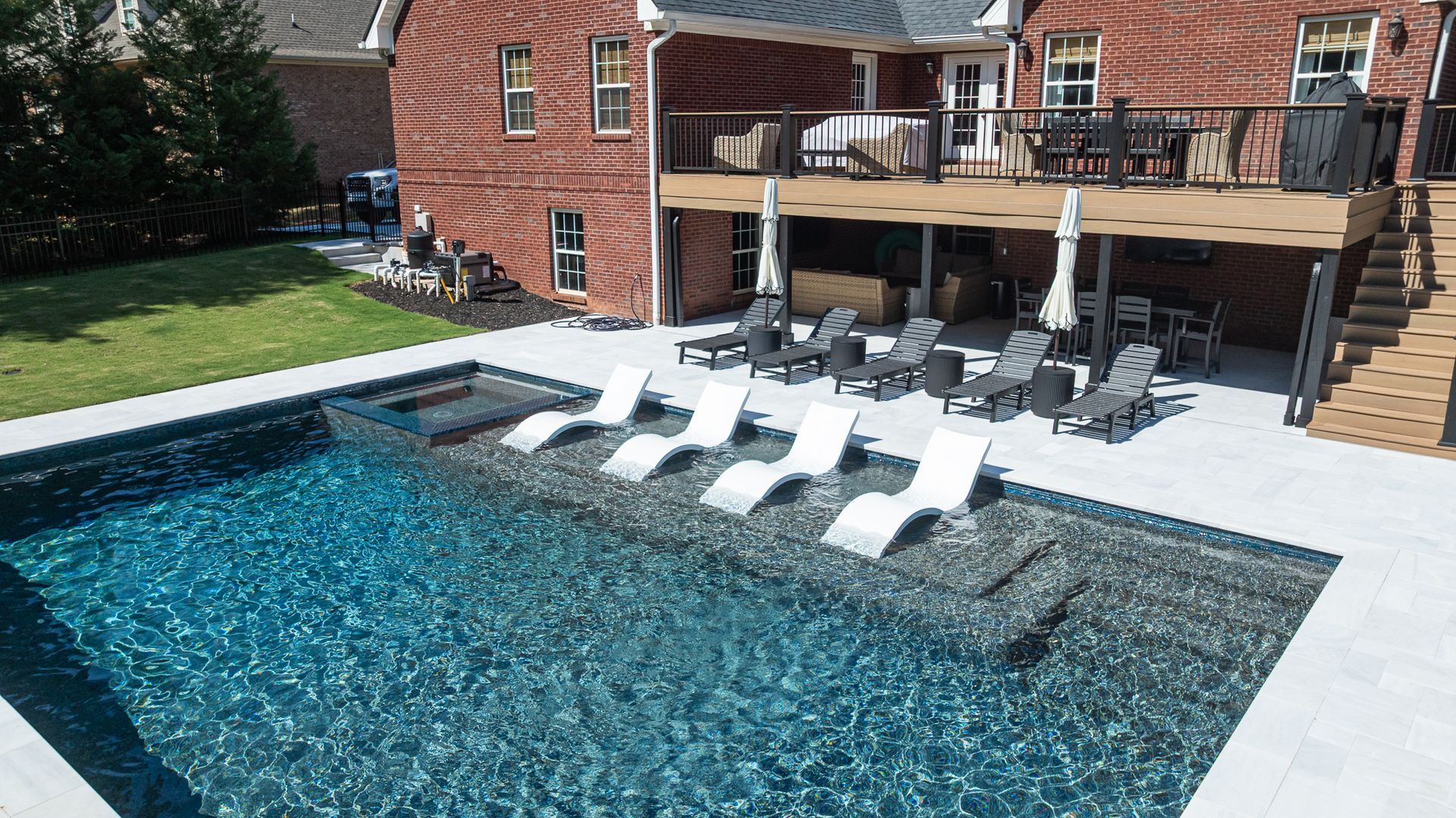 Backyard pool with white lounge chairs and a deck beside a brick house