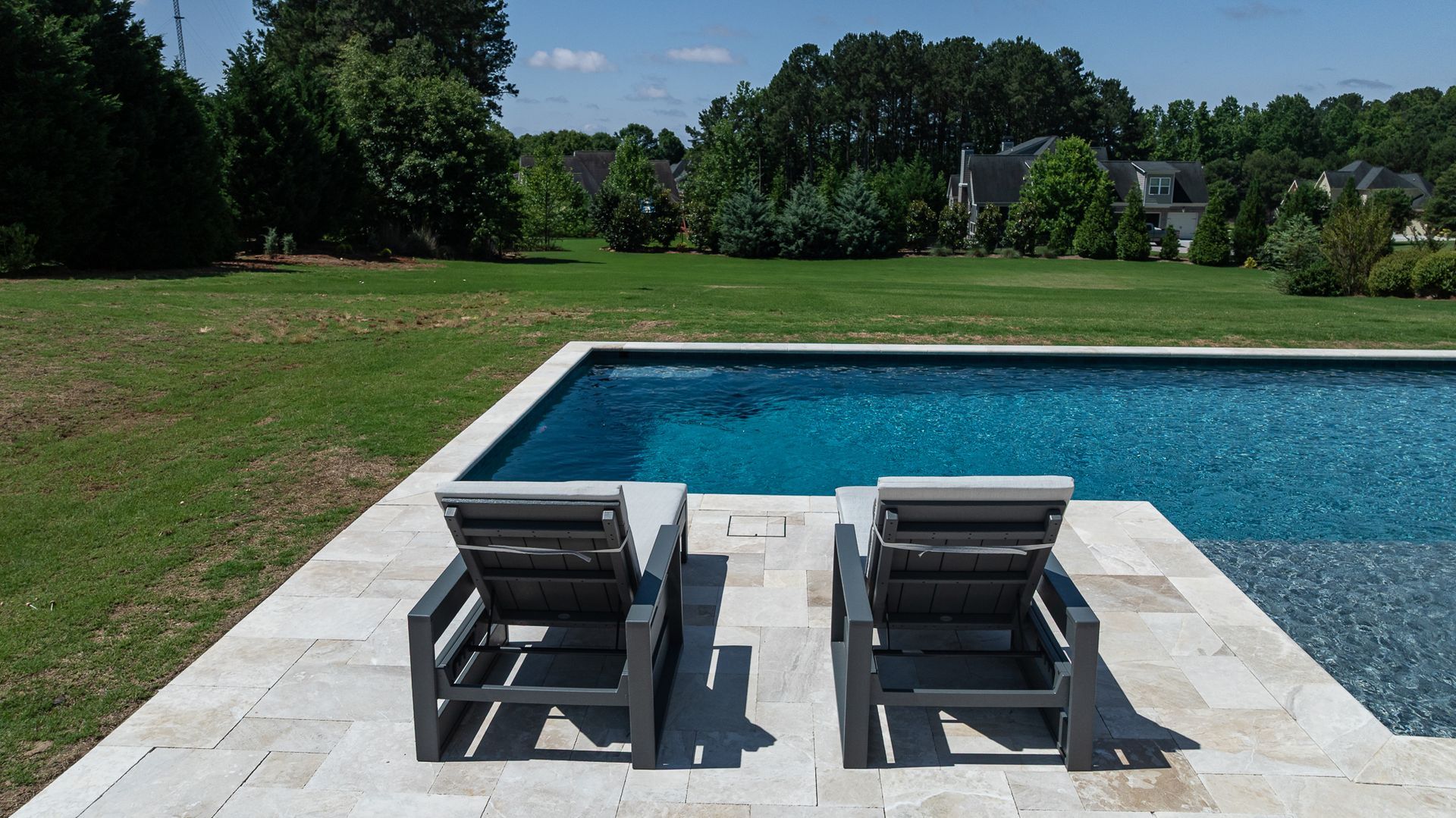 Two gray lounge chairs on a stone patio beside a blue swimming pool with a grassy yard and trees beyond