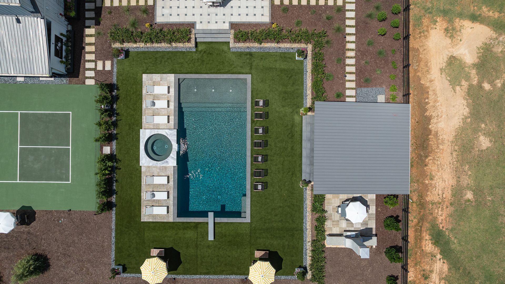 Aerial view of a backyard with a swimming pool, hot tub, lawn, patio, and tennis court nearby