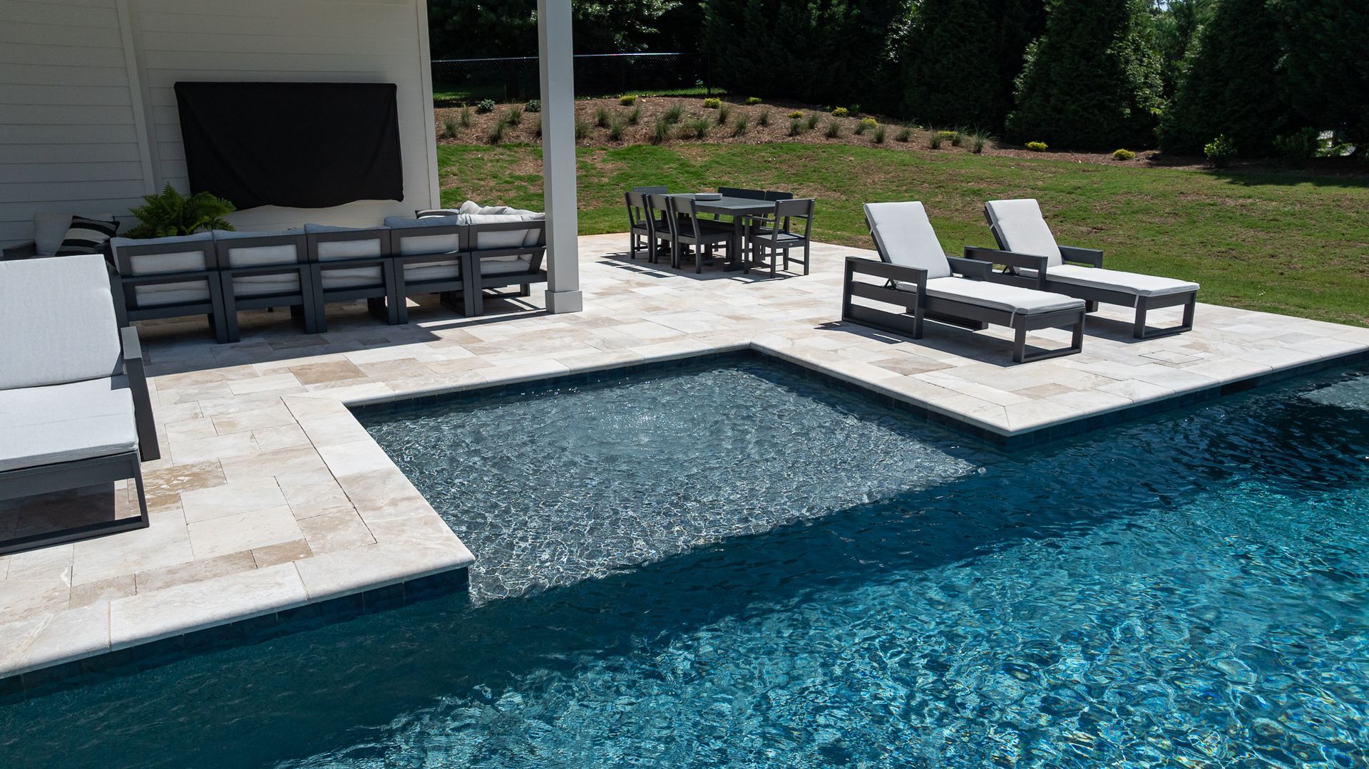 Luxury poolside patio with lounge chairs, tables, and a shallow in-pool ledge beside blue water