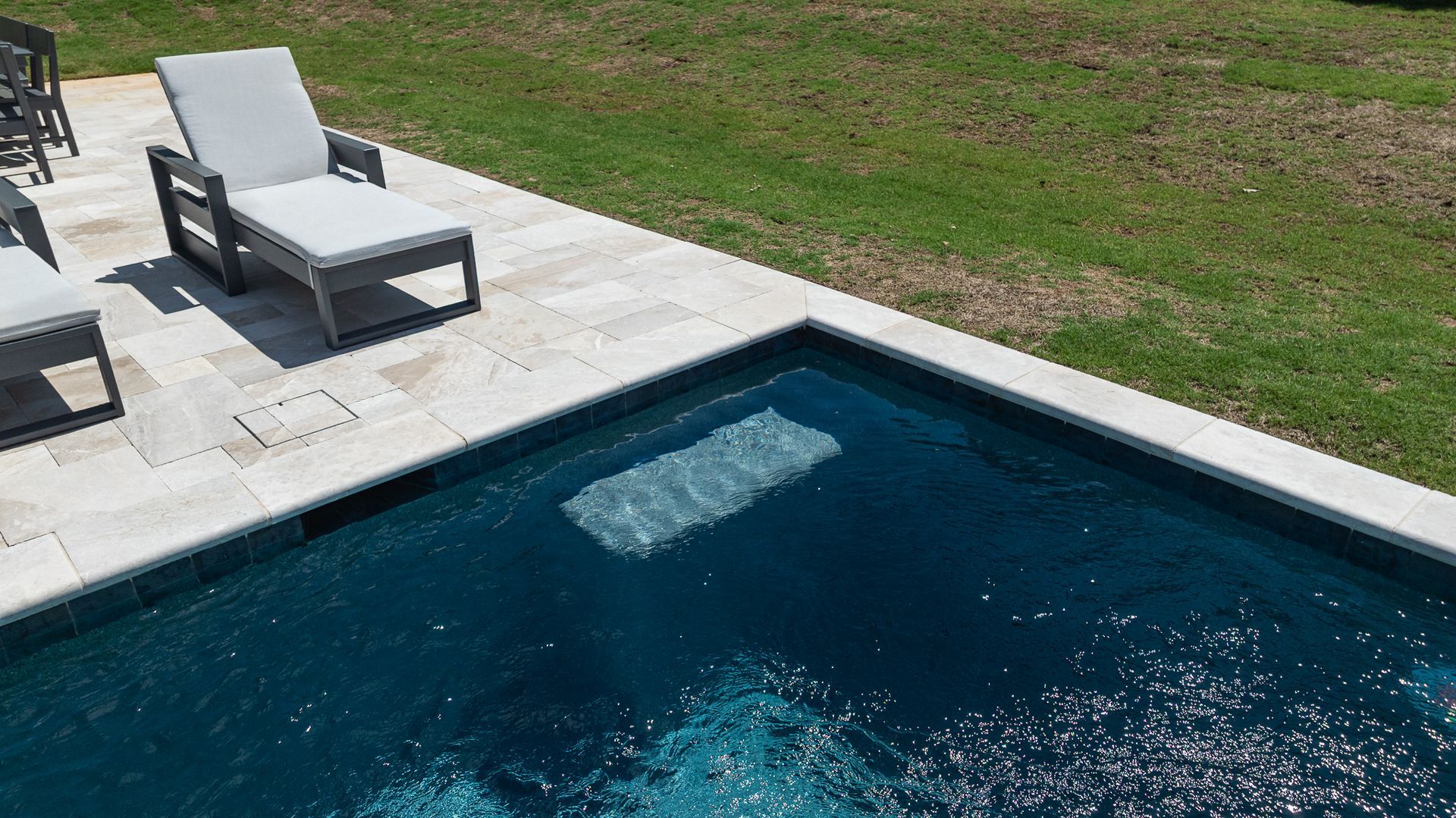 Backyard pool beside a stone patio with two lounge chairs on a grassy lawn.
