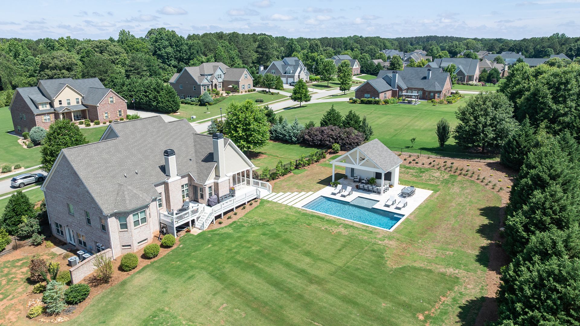 Aerial view of a large suburban home with a pool, patio, and manicured lawn in a wooded neighborhood