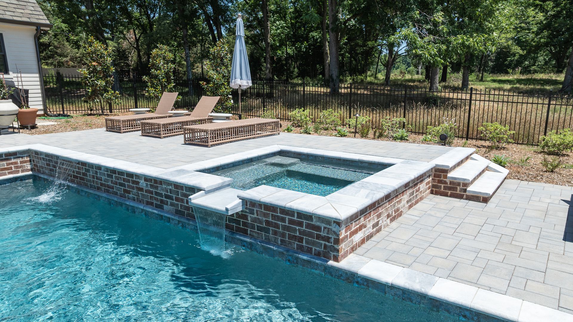 Backyard pool with raised spa, brick surround, lounge chairs, and trees beyond the fence