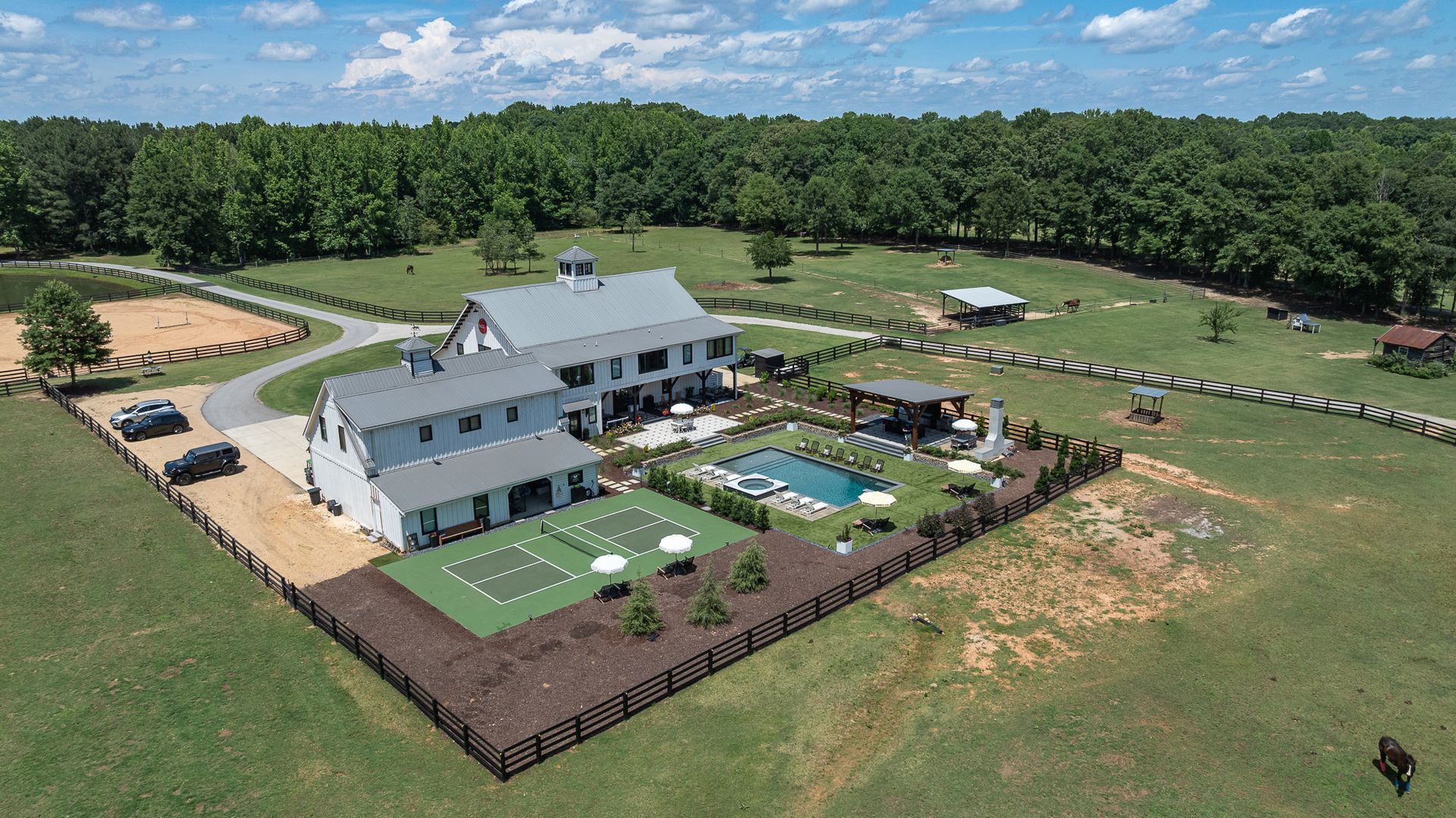 Aerial view of a large gray house with a fenced yard, pool, patio, and tennis court in a green rural landscape