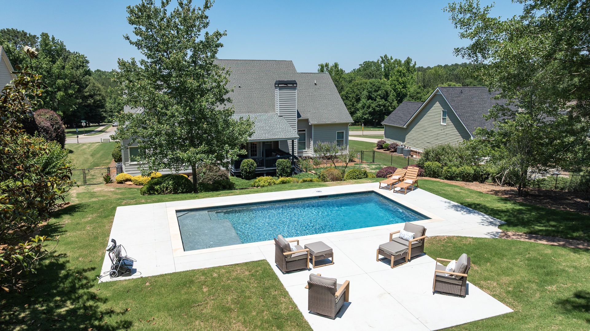Backyard with a rectangular pool, white patio, and outdoor seating beside modern houses and trees