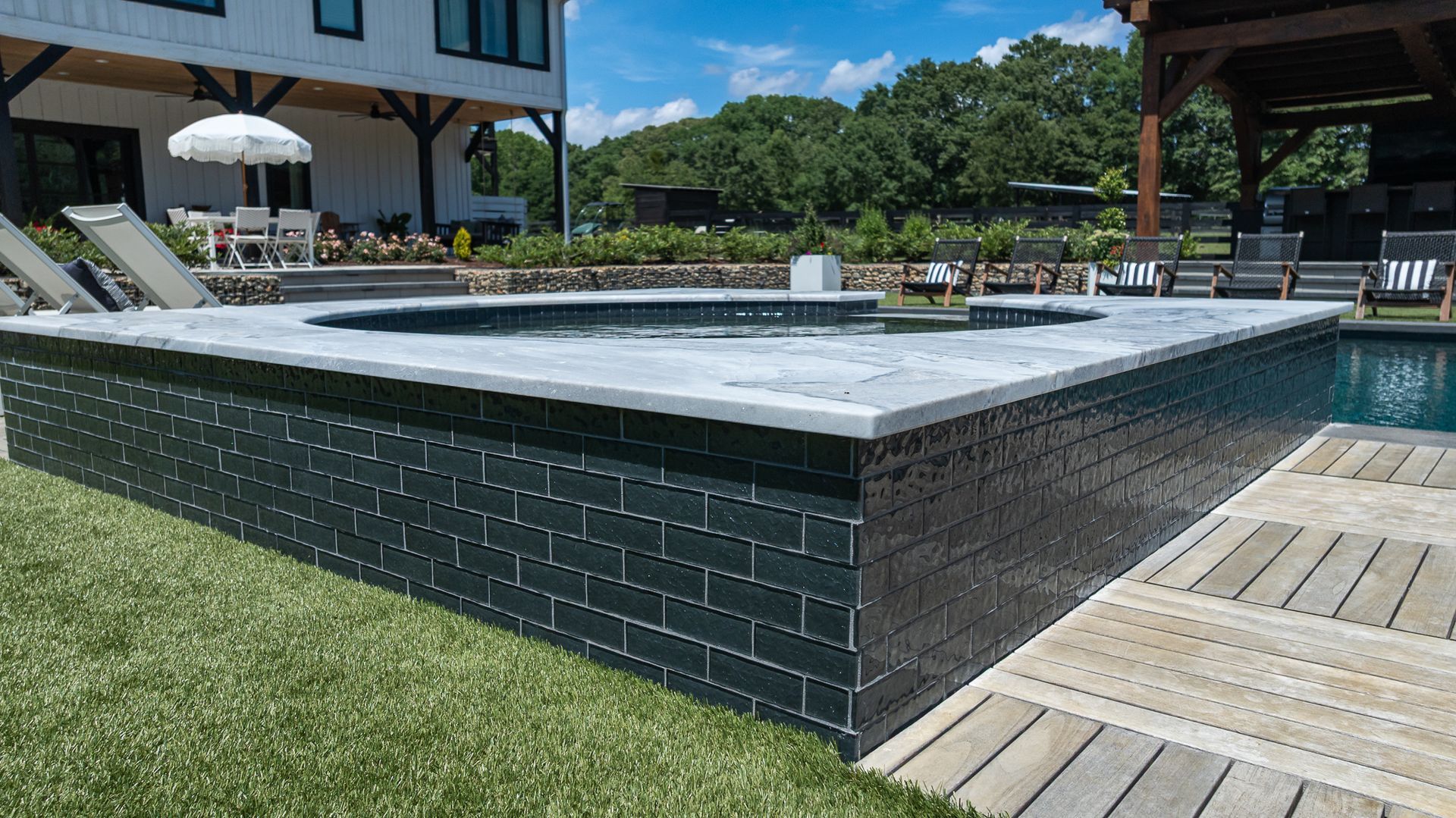 Outdoor swimming pool with dark-tiled raised edge, wooden deck, and modern house in the background