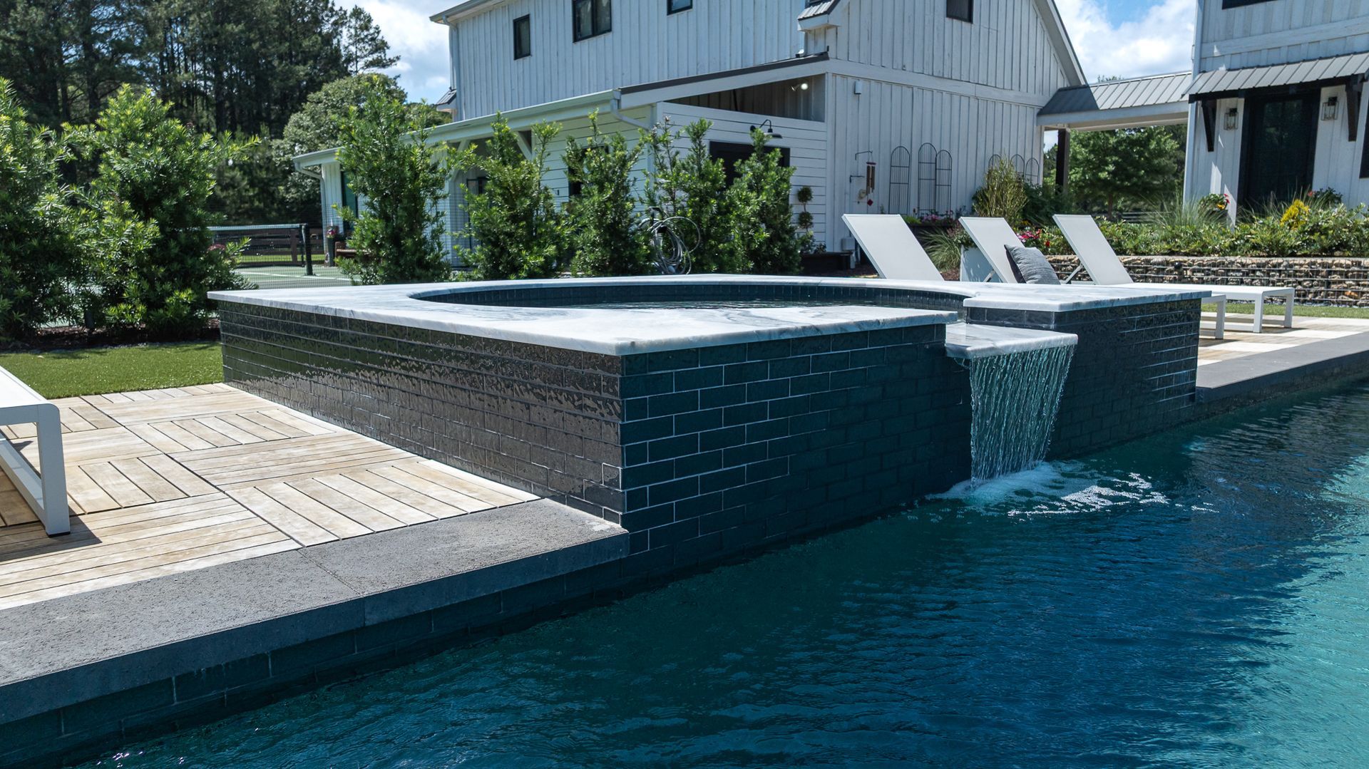 Modern backyard pool with dark tiled edge, lounge chairs, and a white house in the background