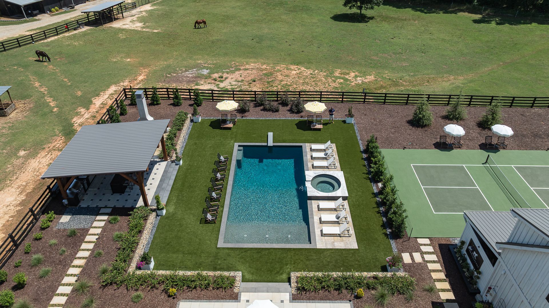 Aerial view of a resort pool, tennis court, and landscaped patio with lounge chairs and umbrellas.