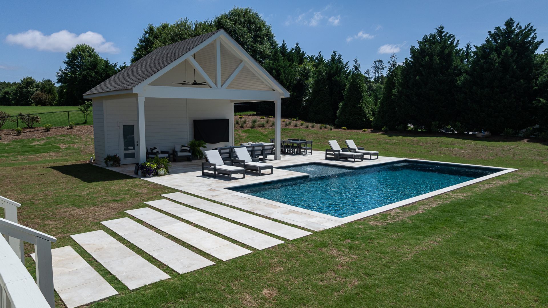 Backyard pool with white lounge chairs, modern pool house, and stone stepping path in a grassy yard.