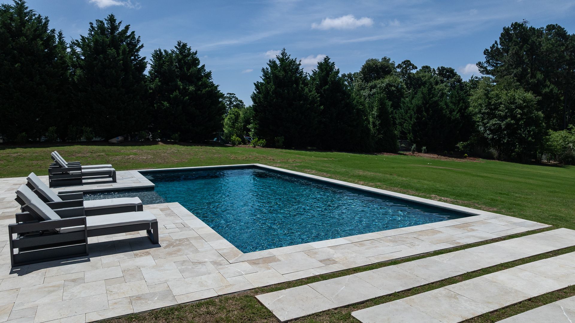 Backyard swimming pool with lounge chairs, stone patio, and manicured lawn under a blue sky