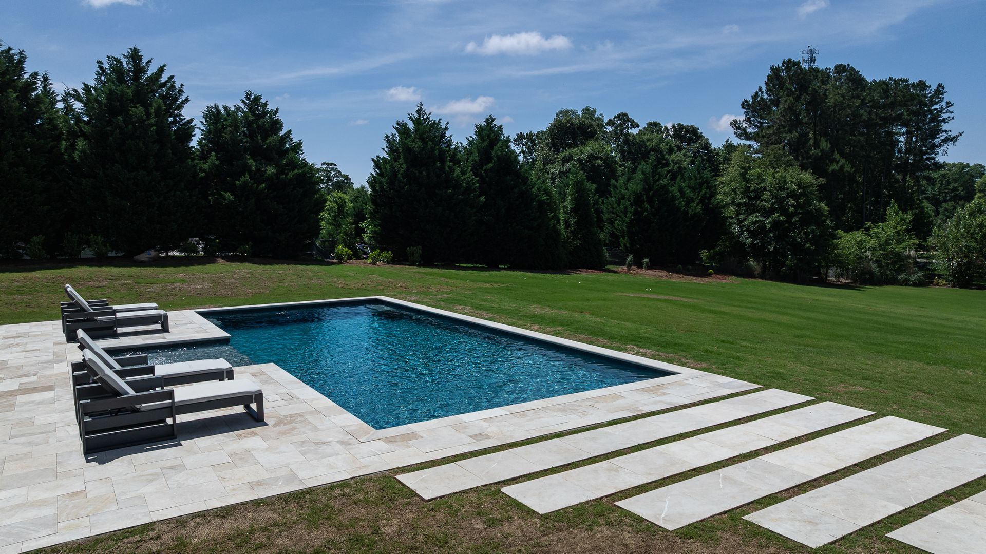 Modern backyard pool with lounge chairs, stepping stones, and a grassy lawn under a blue sky