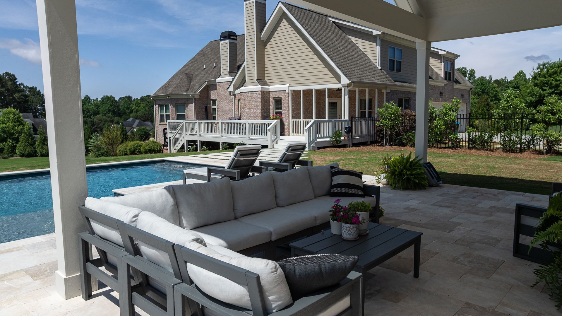 Covered patio with gray outdoor sofas beside a pool and a beige house in the background
