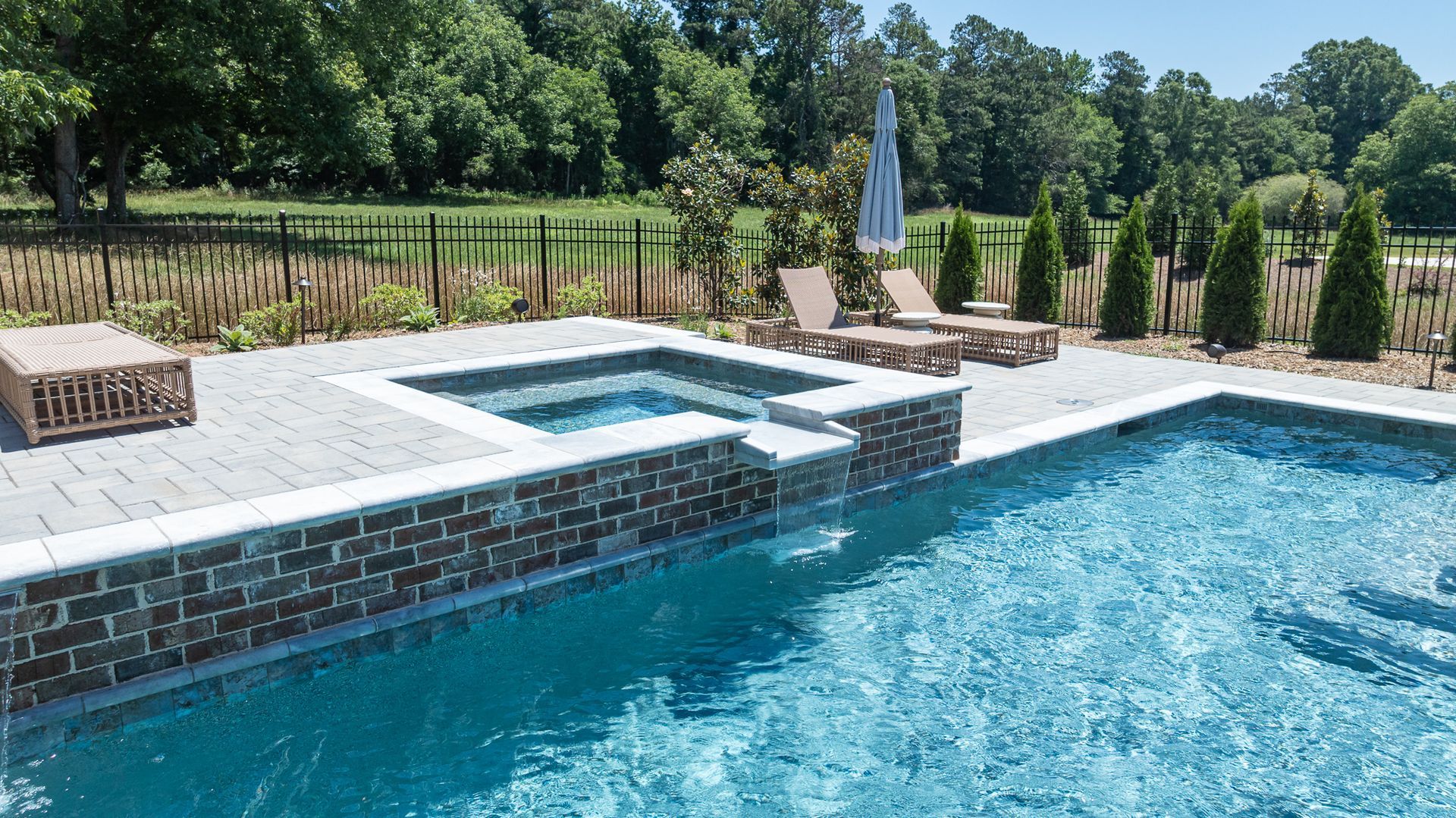 Backyard pool with stone hot tub, blue water, lounge chairs, and green lawn under a sunny sky