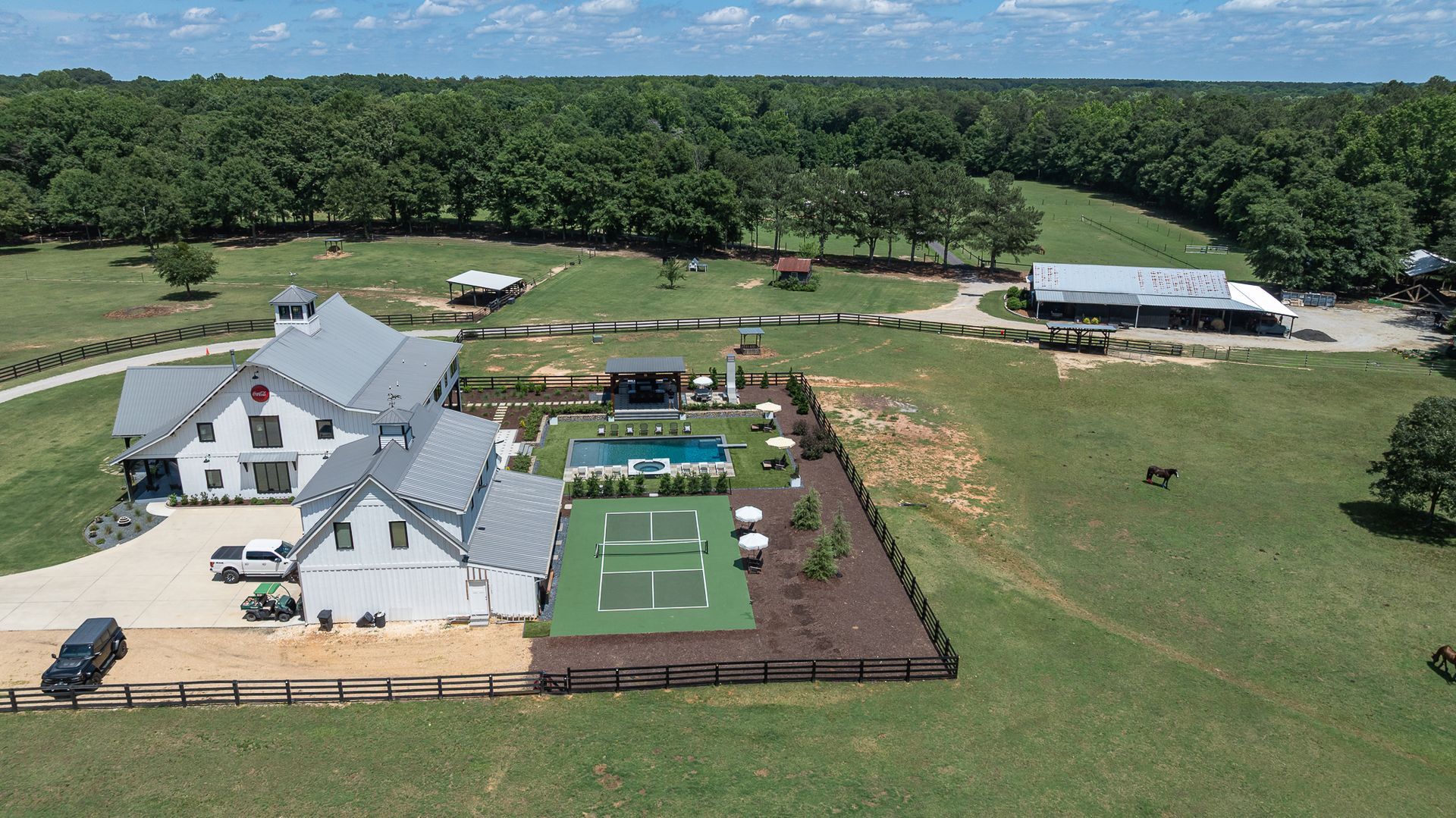 Aerial view of a large white house, lawn, and fenced tennis courts beside a tree-lined field