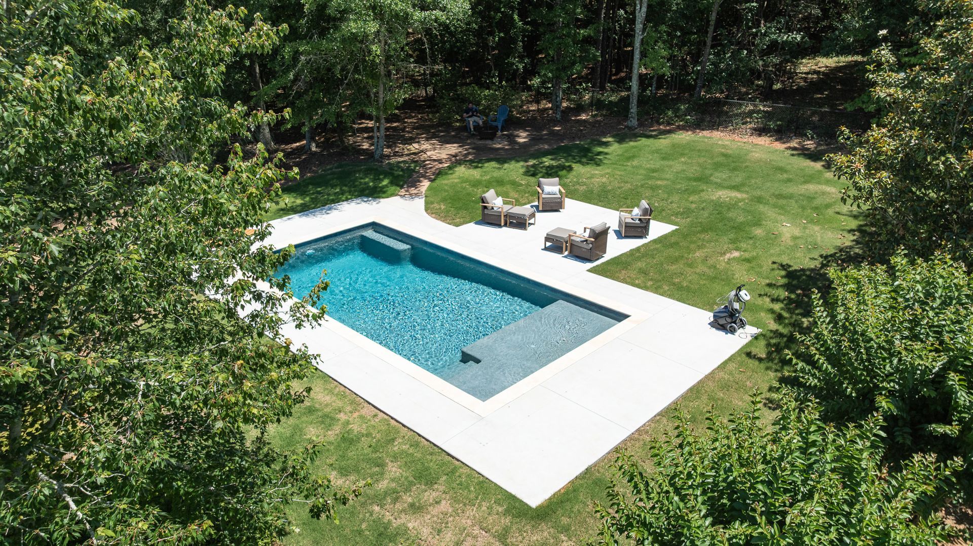 Aerial view of a small backyard pool with white decking, lounge chairs, and surrounding greenery.
