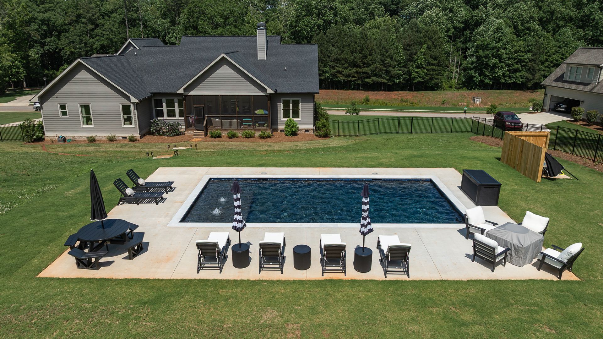 Backyard pool with white lounge chairs, dark umbrellas, and a gray house in the background.