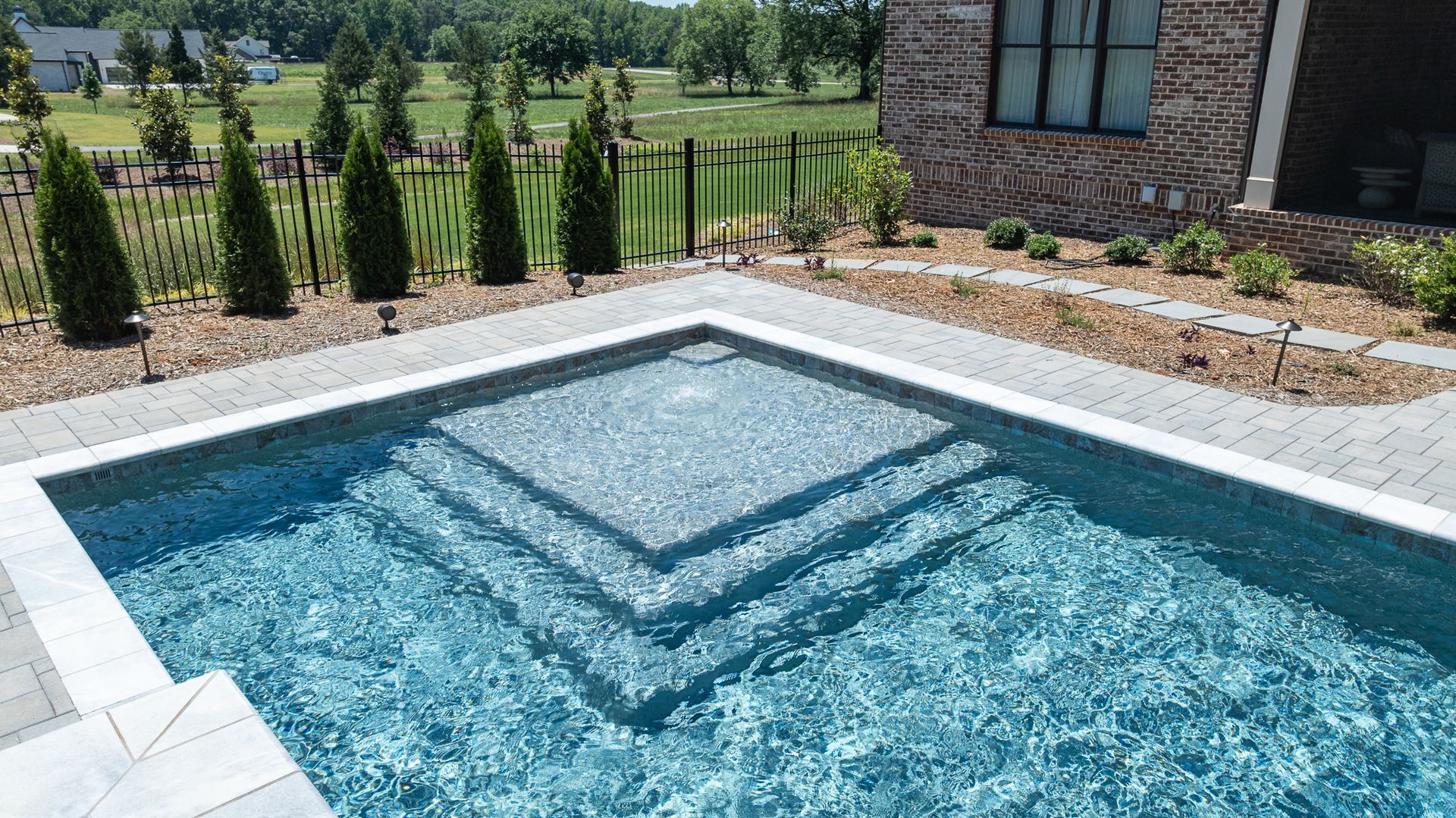 Rectangular backyard pool with blue water and geometric steps beside a stone house and landscaped garden