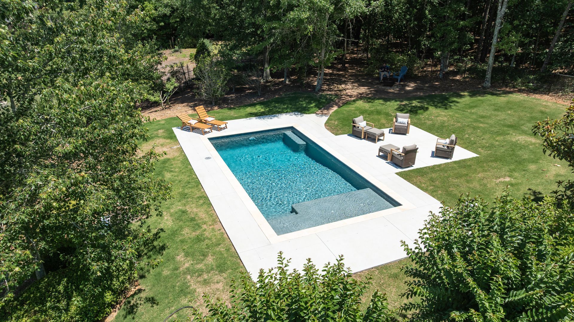 Aerial view of a rectangular backyard pool with white deck chairs and green lawn surrounded by trees