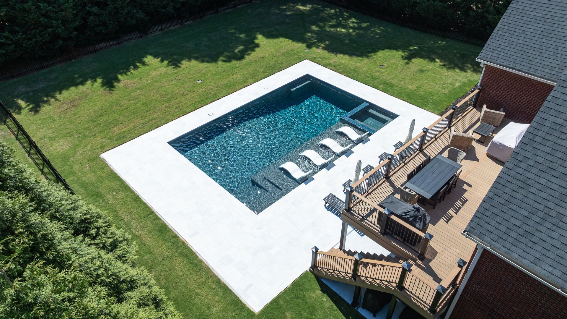 Backyard pool with lounge chairs beside a wooden deck and a brick house, surrounded by green lawn.