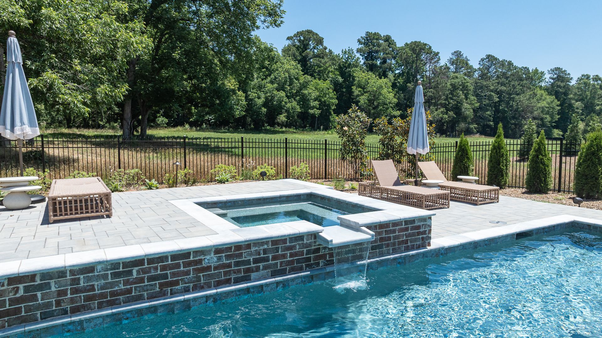 Backyard pool with stone deck, lounge chairs, umbrellas, and a scenic wooded backdrop