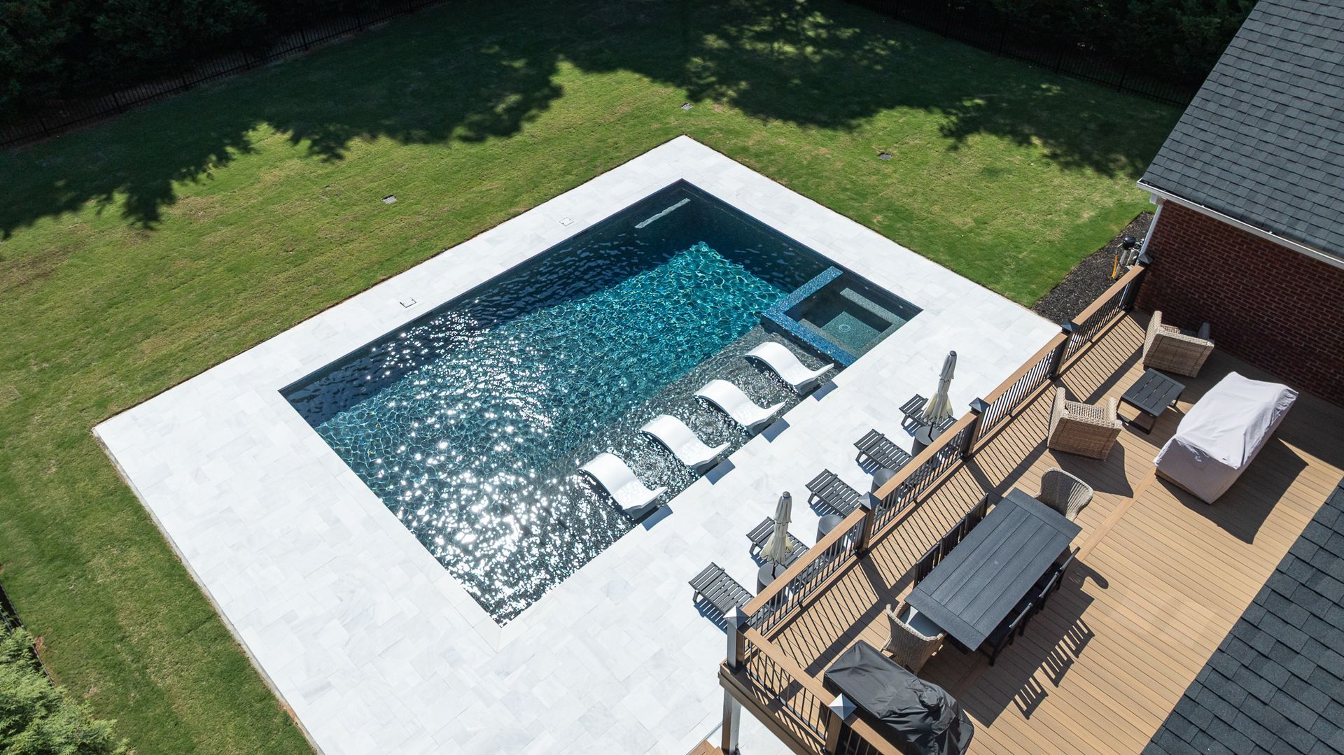 Aerial view of a backyard pool with loungers on a wooden deck and green lawn nearby