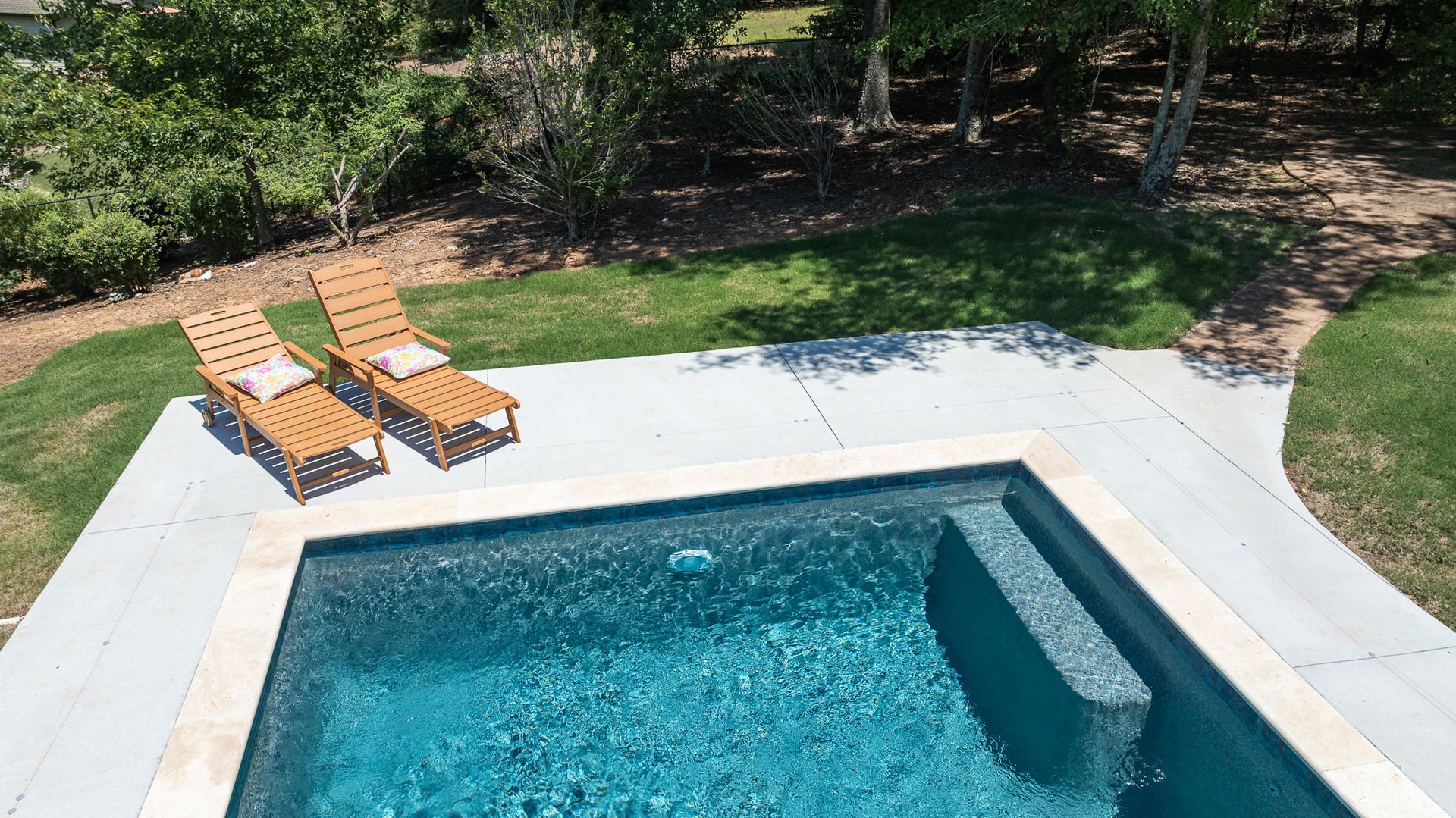 Backyard swimming pool with two wooden lounge chairs on the patio and trees in the background