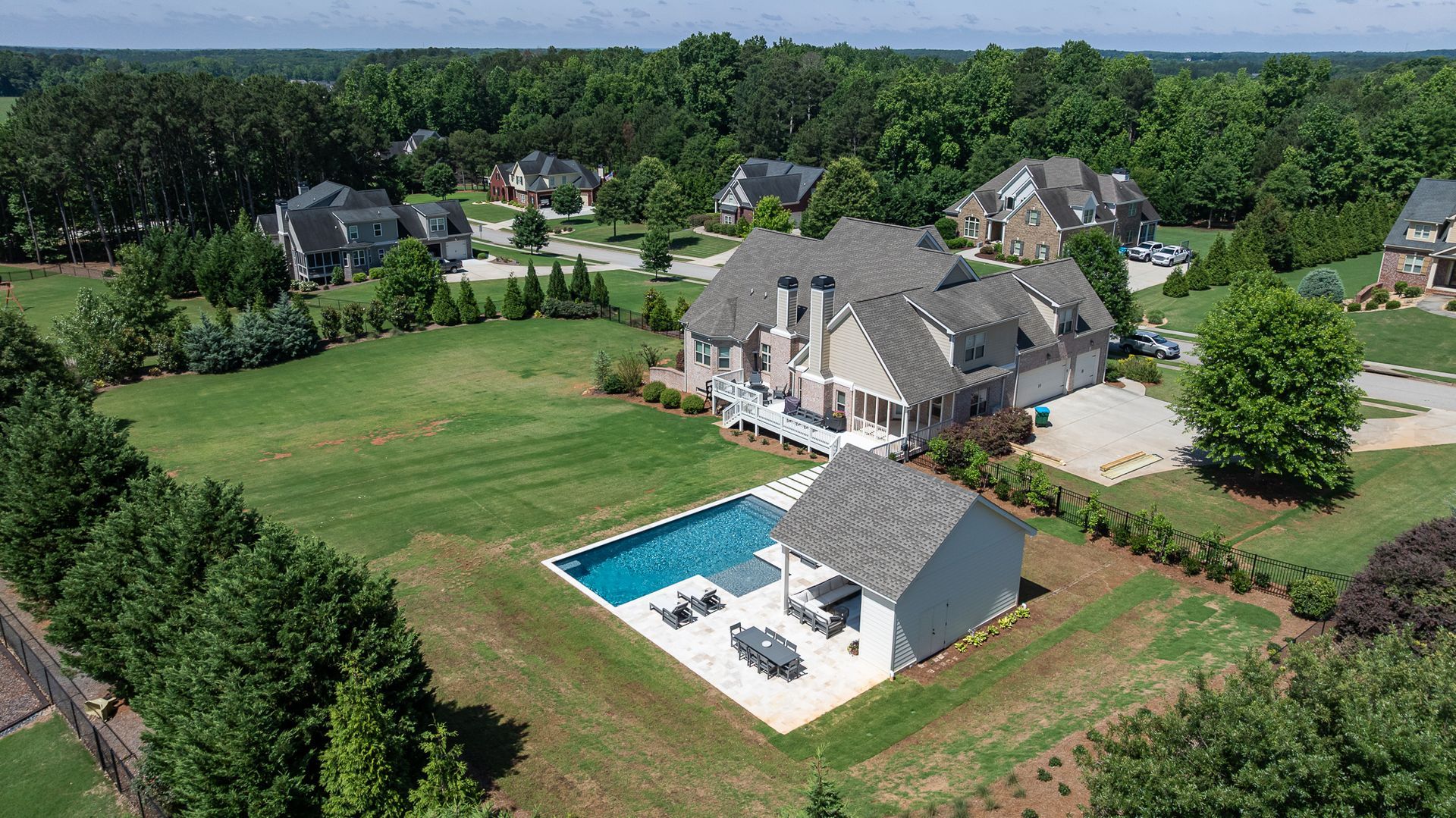 Aerial view of a suburban home with a backyard pool, patio, and surrounding green lawns and trees