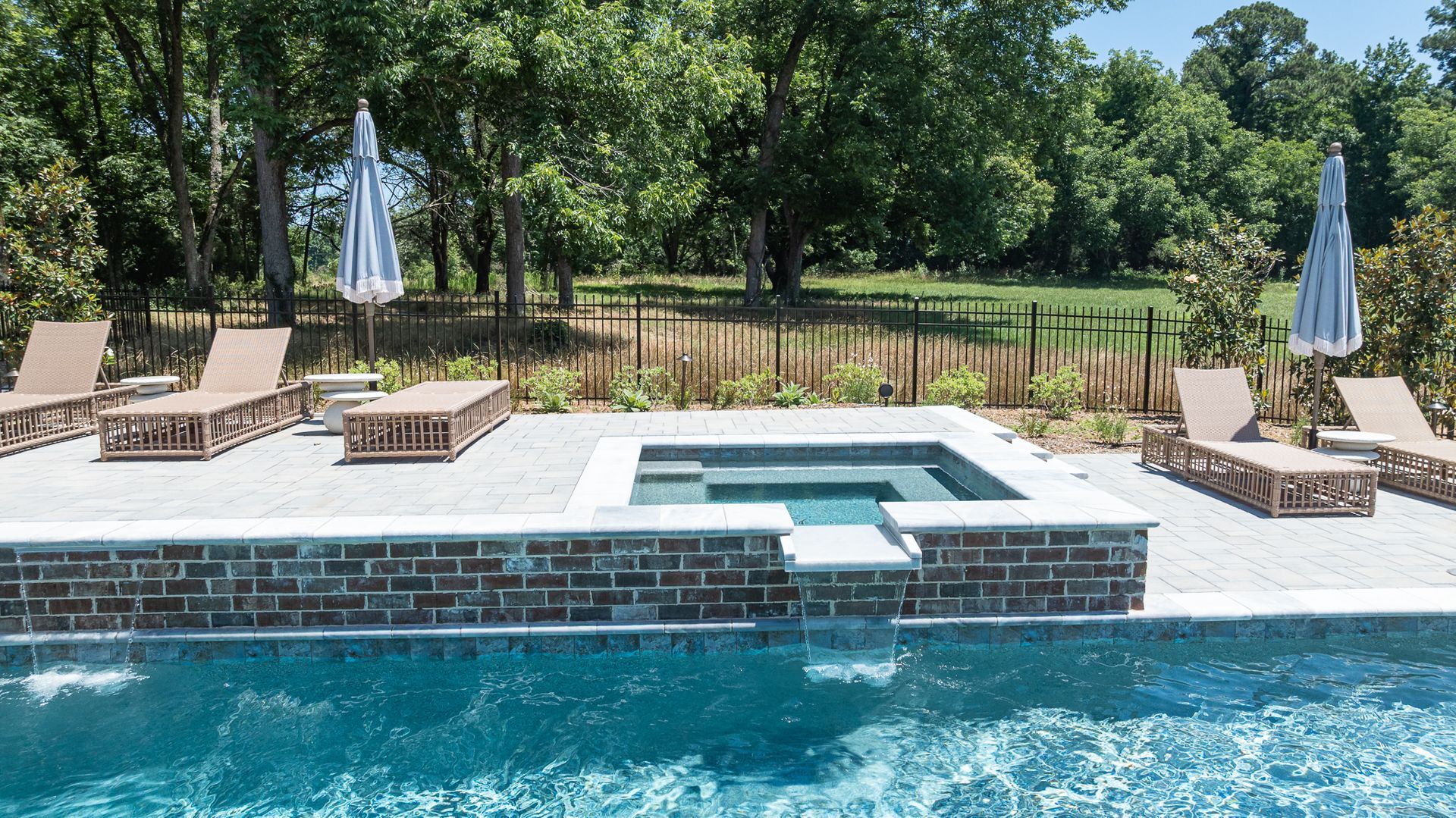 Backyard pool with a raised hot tub, lounge chairs, and umbrellas beside a wooded landscape