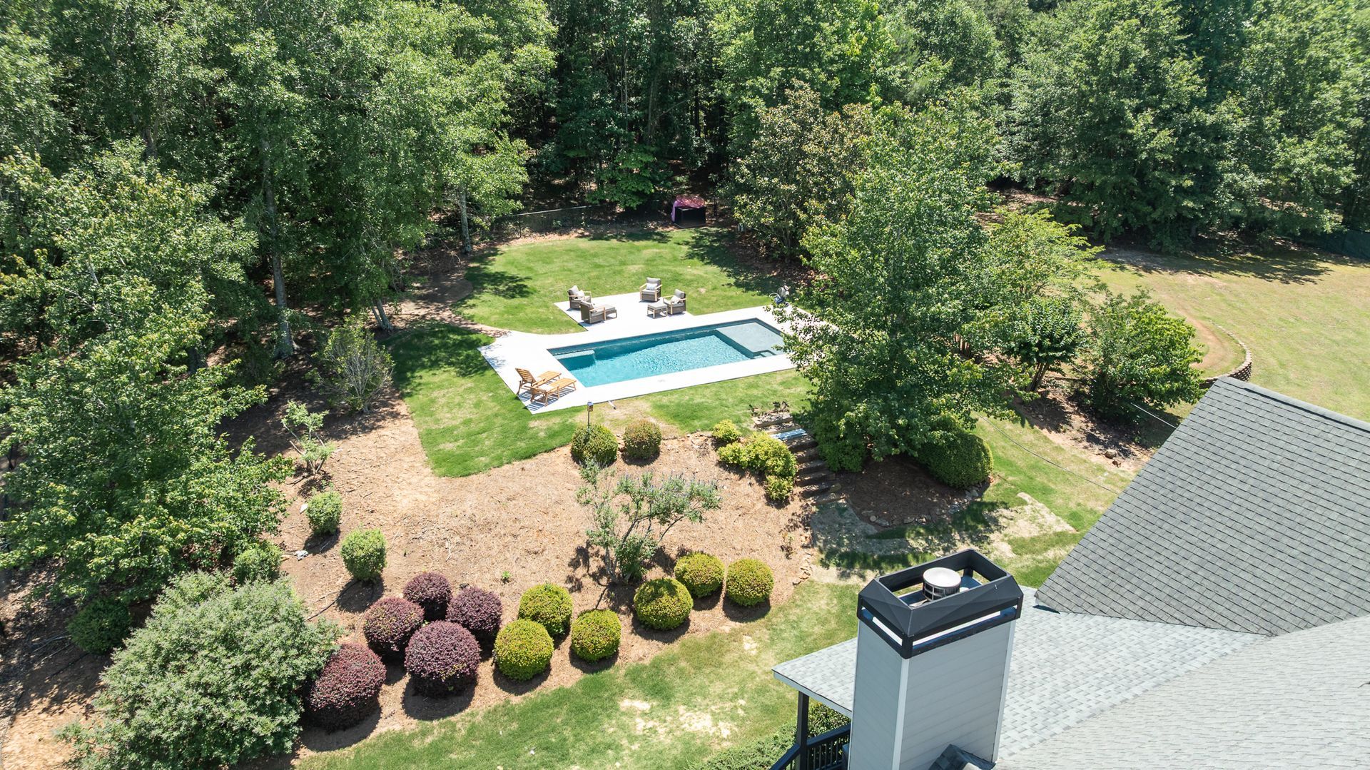 Aerial view of a backyard with a swimming pool, lounge chairs, trees, and a house roof.