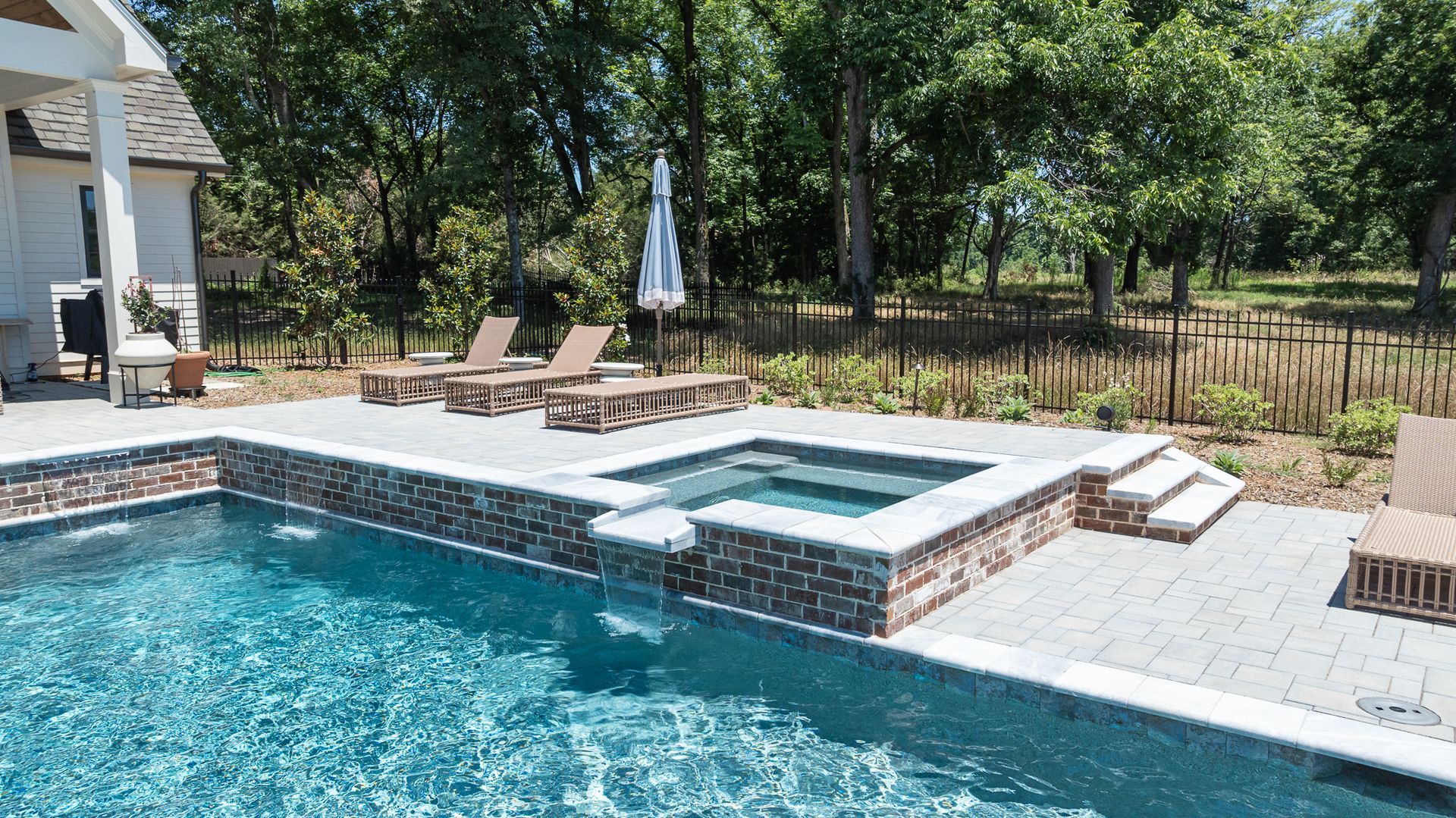 Backyard patio with a swimming pool, raised hot tub, lounge chairs, and trees in the background