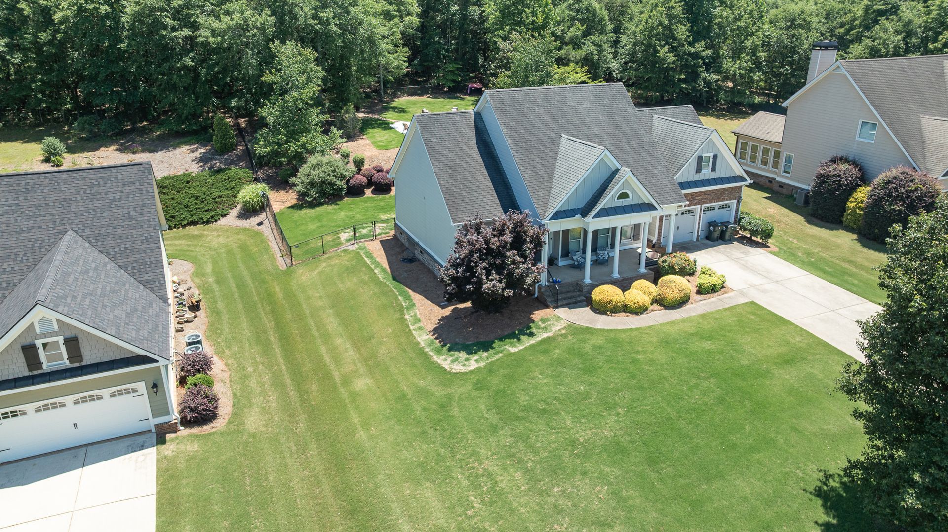 Aerial view of a suburban house with a large green lawn, driveway, and neighboring homes in a wooded neighborhood