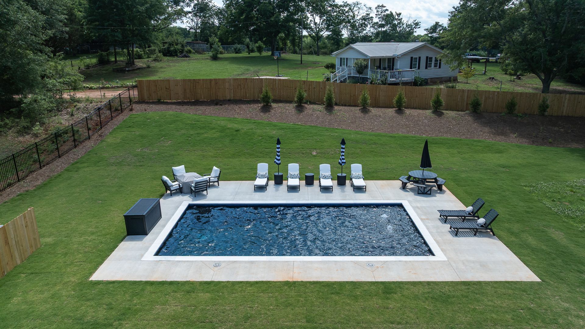 Backyard with a rectangular in-ground pool, patio chairs, and a covered seating area on a green lawn.