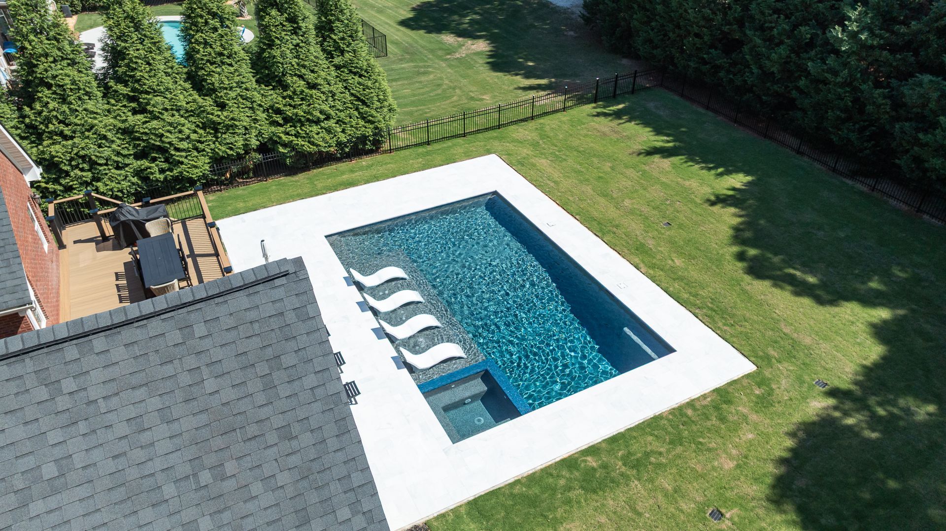 Aerial view of a backyard pool with four white lounge chairs beside a green lawn and trees.