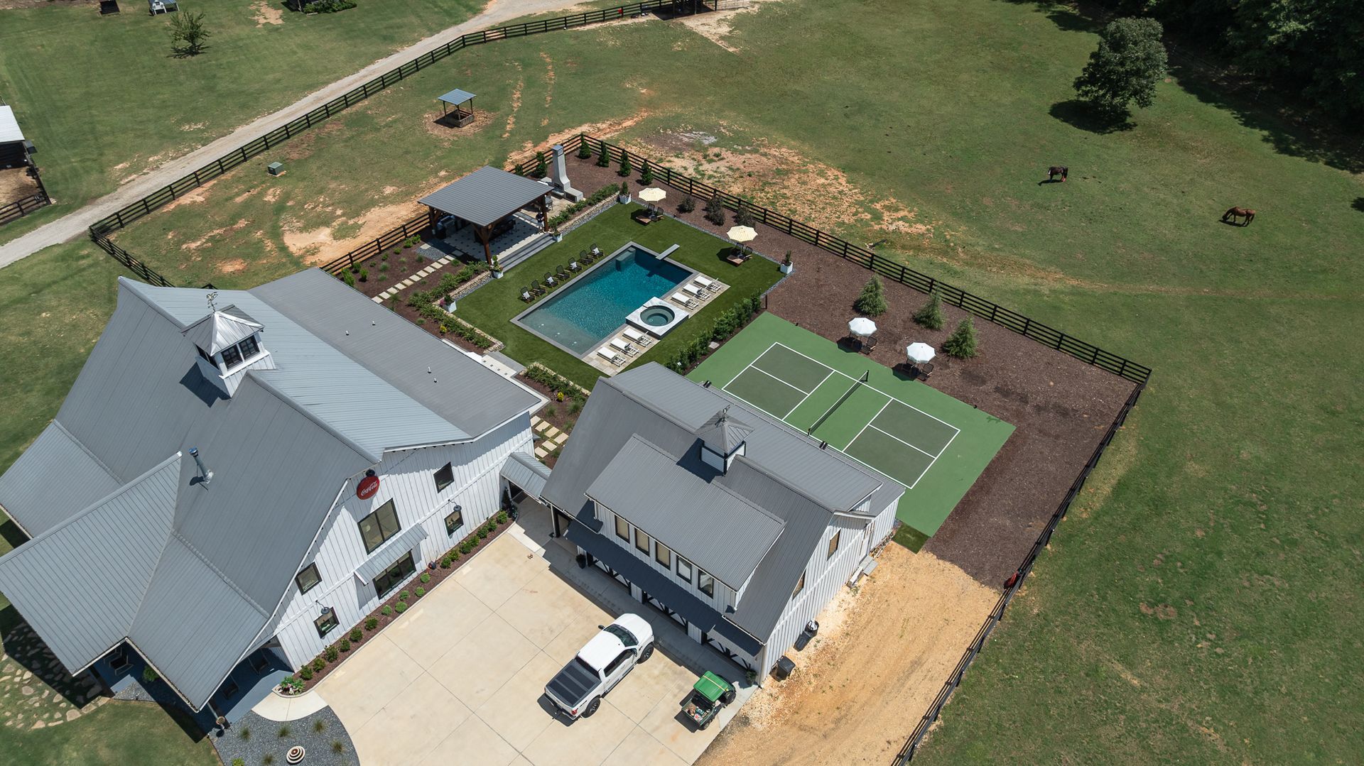 Aerial view of a large white house and gray outbuilding with a pool, patio, and surrounding green lawn.