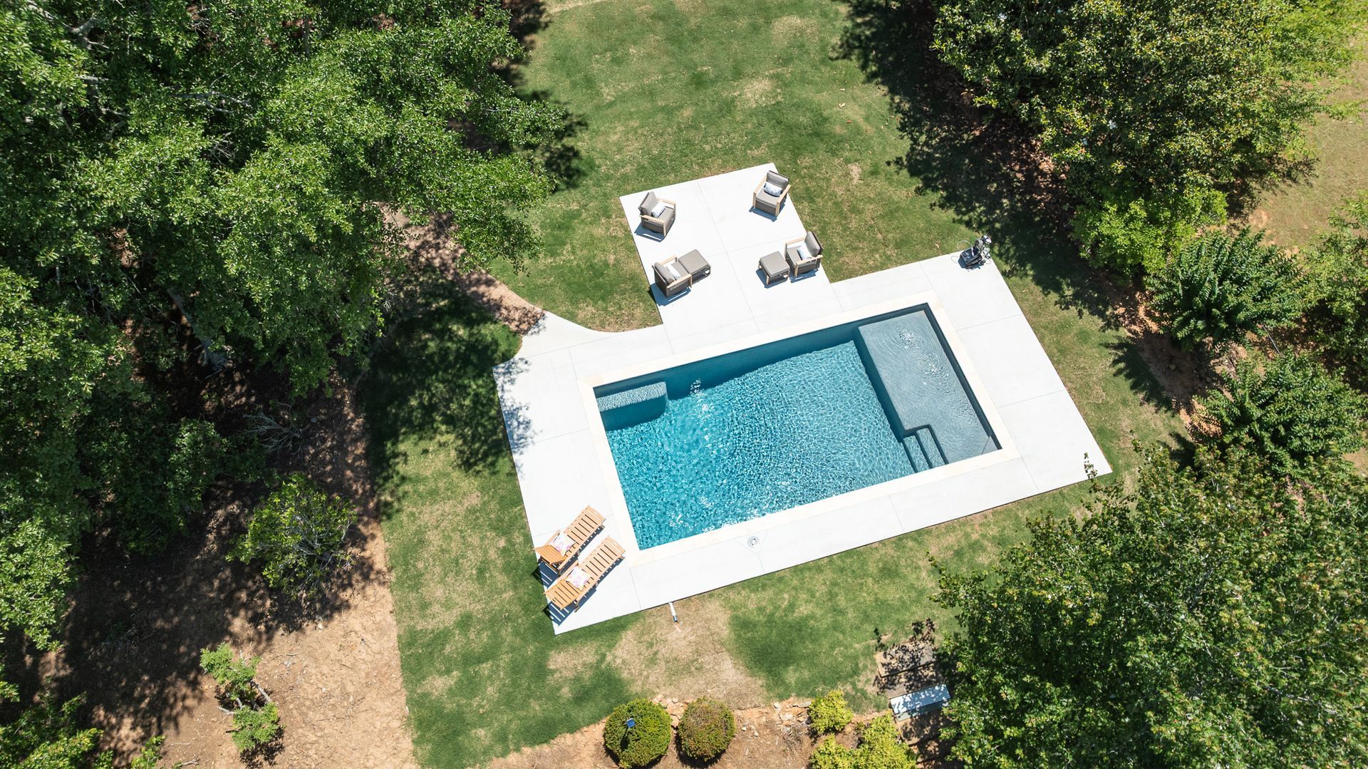 Aerial view of a backyard pool with white patio chairs and lush green trees around it.