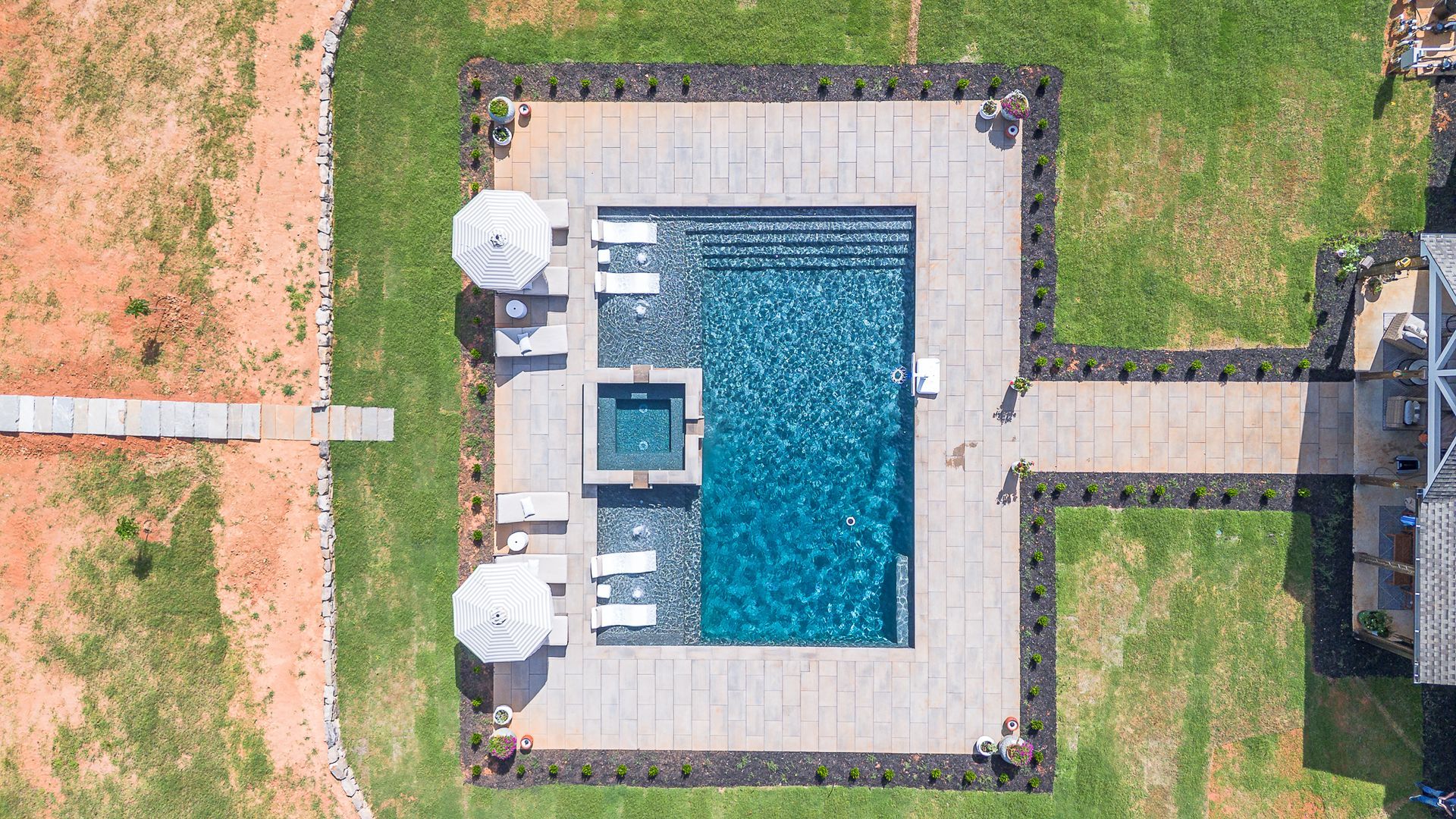 Aerial view of a rectangular pool surrounded by stone patio, landscaping, and lawn. Two lounge chairs and umbrellas next to pool.