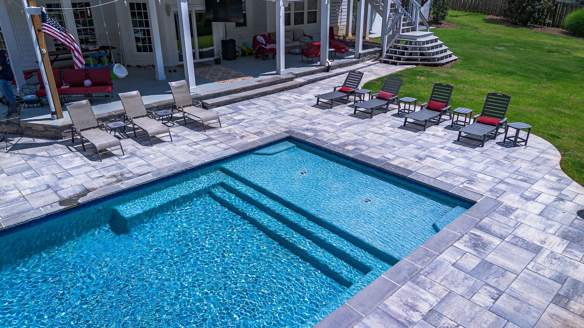 Pool with blue water, steps, and surrounding patio with lounge chairs. Exterior shot with a house in background.