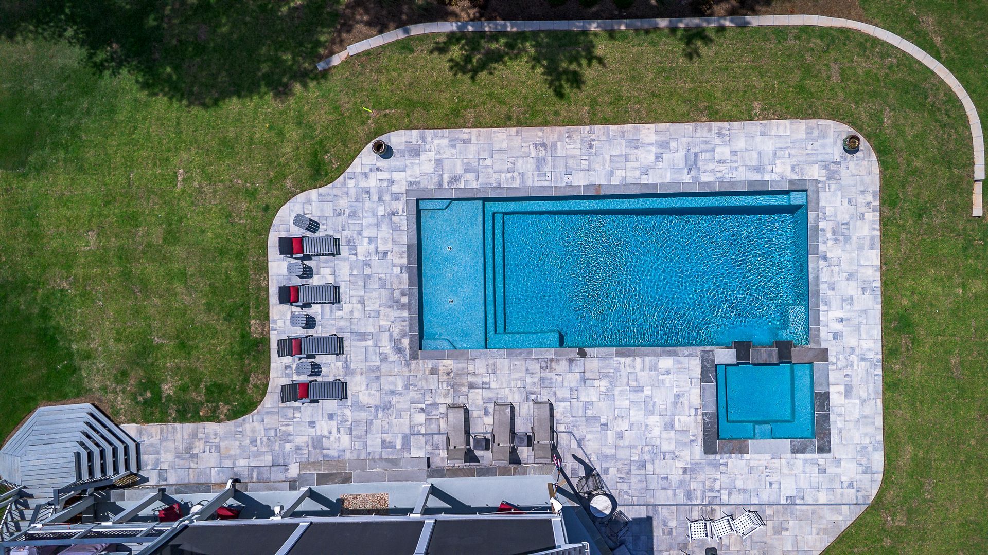 Overhead view of a rectangular pool with a hot tub, surrounded by stone patio and green grass.