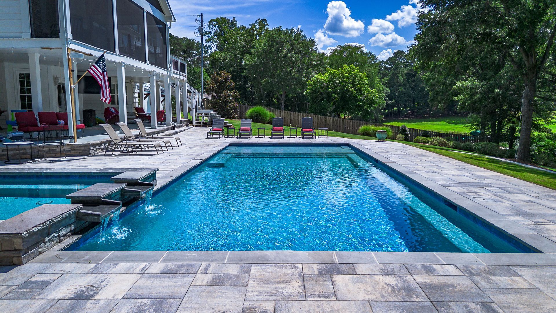 A large rectangular pool with blue water, surrounded by a stone patio and a white house on a sunny day.