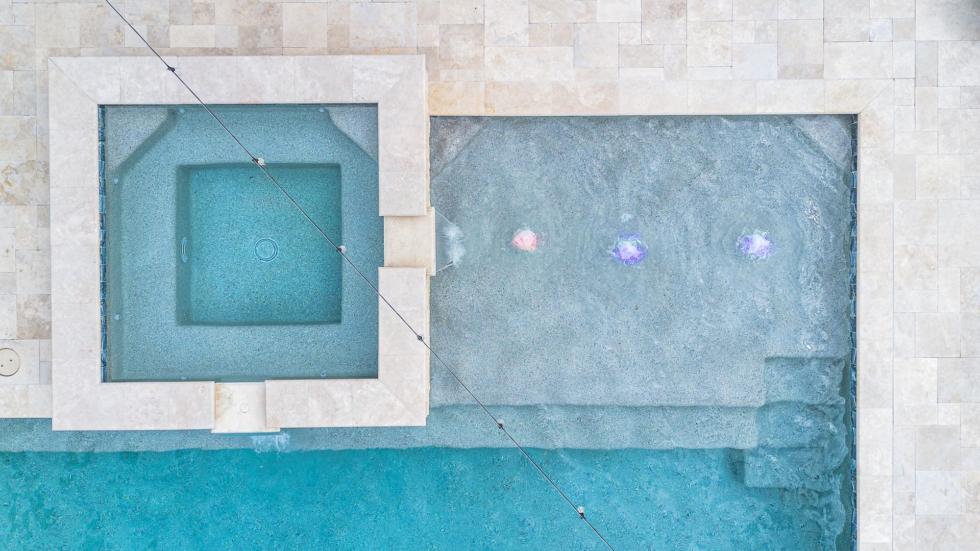 Overhead view of a swimming pool and spa with turquoise water, light-colored stone edging, and submerged lights.