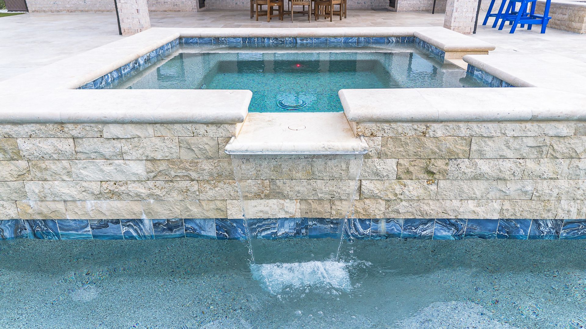 Small pool with waterfall feature, surrounded by stone and blue tile.