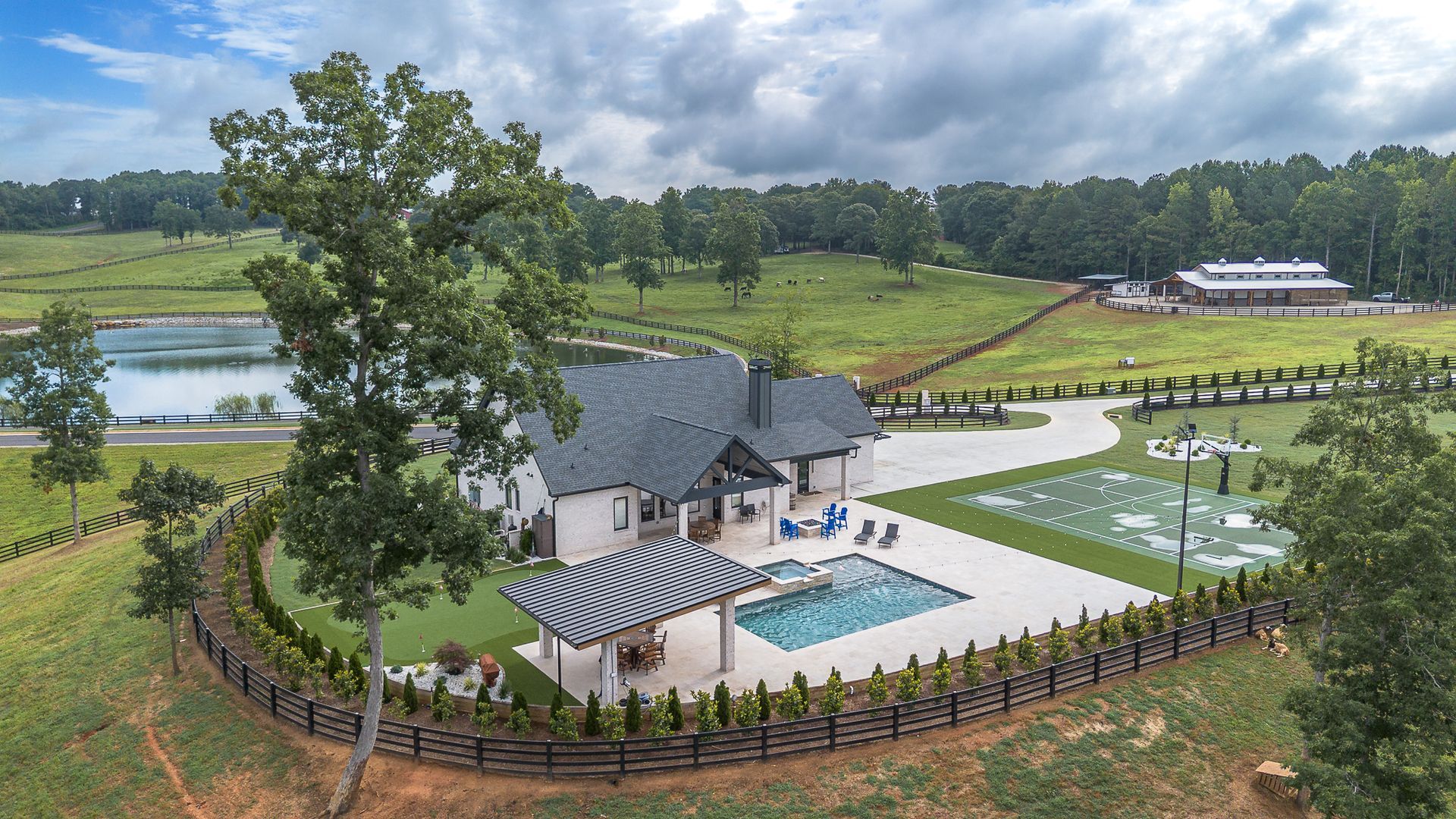 Spacious house with pool, patio, and basketball court. Fields, trees, and lake surround it. Overcast sky.