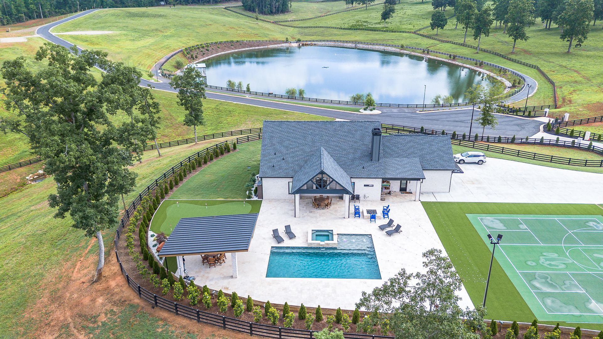 Aerial view of a modern house with pool, tennis court, and large pond in a rural setting.