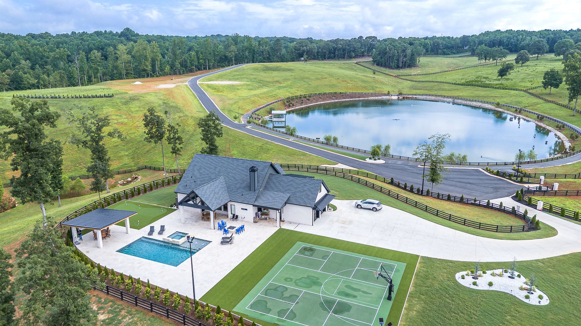 Luxury home with pool, sports court, and lake, surrounded by trees and green fields.