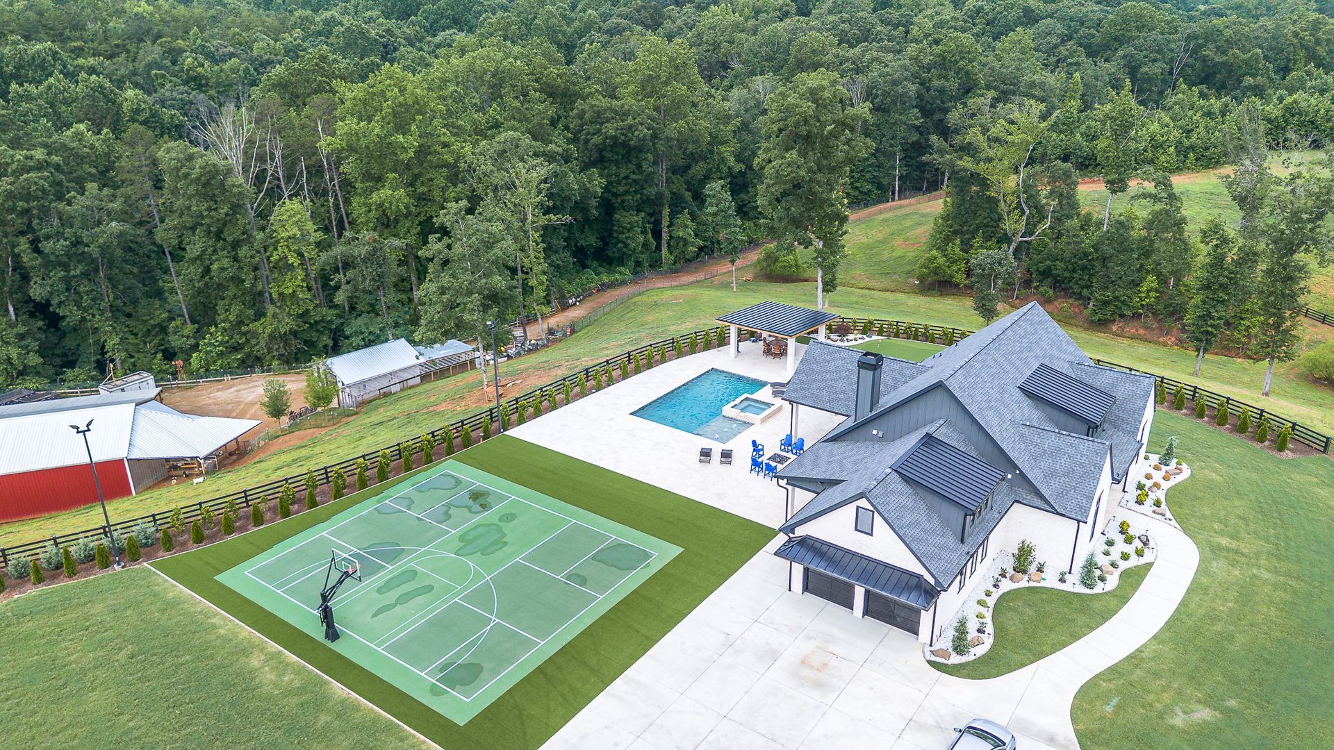 Aerial view of a luxury home with a pool, basketball court, and forest backdrop.
