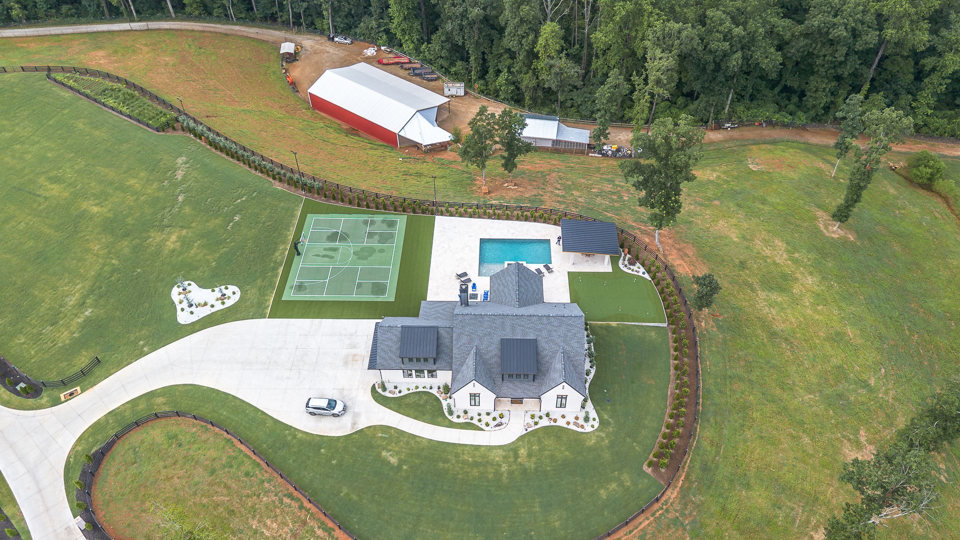 Aerial view of a large house with pool, tennis court, and red barn on a grassy landscape.